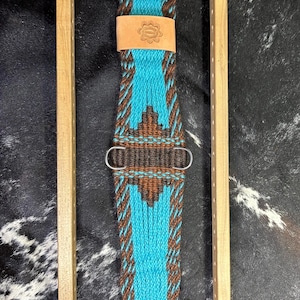 May include: A turquoise and brown woven horse cinch with a leather patch featuring a floral design. The cinch has silver-colored metal buckles and hardware. The design includes a central turquoise panel with brown geometric patterns and borders.