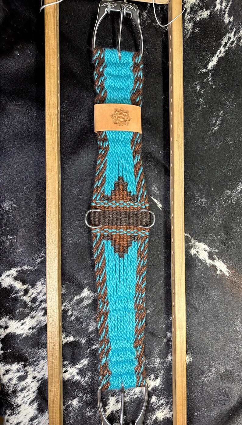 May include: A turquoise and brown woven horse cinch with a leather patch featuring a floral design. The cinch has silver-colored metal buckles and hardware. The design includes a central turquoise panel with brown geometric patterns and borders.