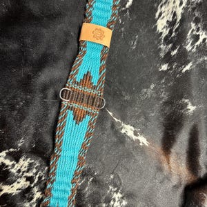 May include: A turquoise and brown woven horse cinch with silver buckles and a leather patch. The cinch features a geometric pattern and is designed for equestrian use. The leather patch has a floral design.