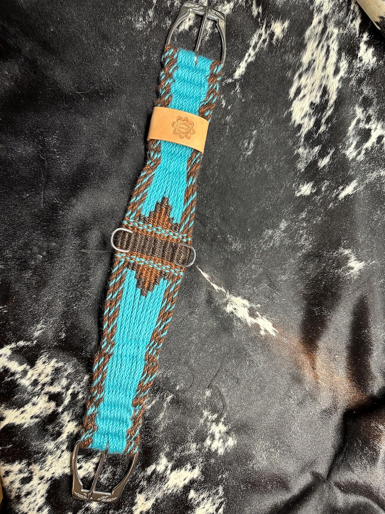 May include: A turquoise and brown woven horse cinch with silver buckles and a leather patch. The cinch features a geometric pattern and is designed for equestrian use. The leather patch has a floral design.