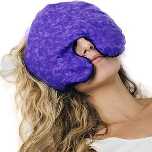 Puede incluir: Una máscara de ojos morada con ribete negro, diseñada para cubrir los ojos y la frente. La máscara está hecha de una tela texturizada. La persona de la imagen tiene el pelo largo y rubio ondulado. La máscara está diseñada para la relajación.