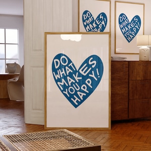 May include: Three framed art prints with a white background and a blue heart design. The heart contains the words "DO WHAT MAKES YOU HAPPY!" in white. The frames are a light brown colour. The prints are displayed in a room with wooden floors.