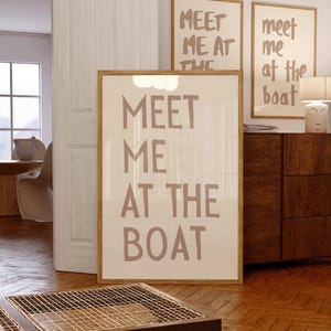May include: A beige framed poster with the text "MEET ME AT THE BOAT" in a handwritten style font. The poster is hanging on a wall in a room with wooden floors.