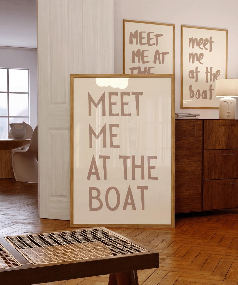 May include: A beige framed poster with the text "MEET ME AT THE BOAT" in a handwritten style font. The poster is hanging on a wall in a room with wooden floors.