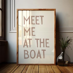 May include: A framed print with the text "MEET ME AT THE BOAT" in a light brown color on a white background. The frame is made of wood and has a light brown finish.