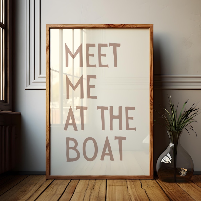 May include: A framed print with the text "MEET ME AT THE BOAT" in a light brown color on a white background. The frame is made of wood and has a light brown finish.