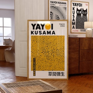 May include: Three framed art prints by Yayoi Kusama. The central print has a yellow dotted pattern and Japanese text. The other two prints feature black and white designs and Japanese text. The prints are displayed in a room with wooden floors and a wooden cabinet.