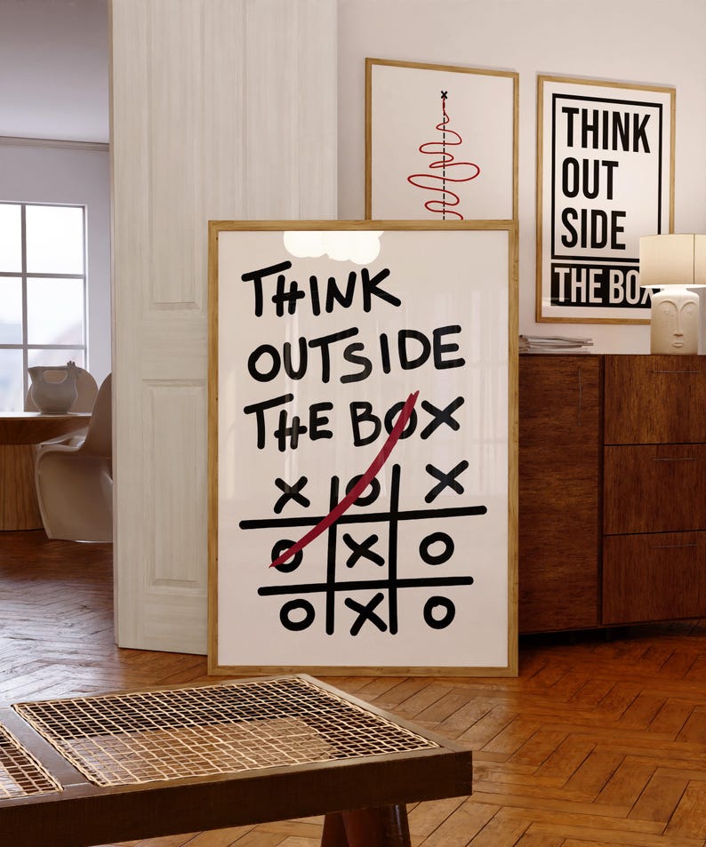 Puede incluir: Un p&oacute;ster enmarcado en blanco con el texto "Think Outside The Box" en marcador negro. Una l&iacute;nea roja se dibuja a trav&eacute;s de un juego de tres en raya en el p&oacute;ster.