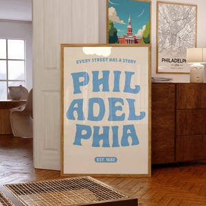 May include: A framed poster with the word "PHILADELPHIA" in large, light blue letters. The poster also includes the text "EVERY STREET HAS A STORY" and "EST. 1682". Two other framed images are visible in the background.