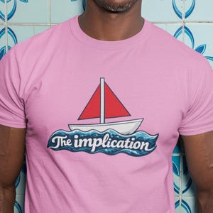 May include: A light pink t-shirt featuring a sailboat with a red sail and the text "The implication" in a blue font. The shirt is worn by a person wearing sunglasses.