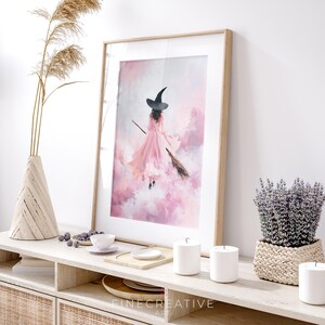 May include: A framed art print featuring a witch in a pink dress and black hat flying on a broomstick through a pink and white cloudy sky. The artwork is displayed on a light-colored wooden console table with decorative items.