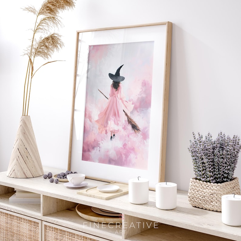 May include: A framed art print featuring a witch in a pink dress and black hat flying on a broomstick through a pink and white cloudy sky. The artwork is displayed on a light-colored wooden console table with decorative items.