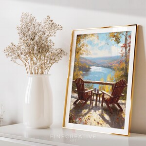 May include: A framed art print featuring a scenic view of a lake and two wooden chairs on a deck. The artwork has a gold frame and depicts an autumn landscape with vibrant colors. A white vase with dried flowers is on the left.