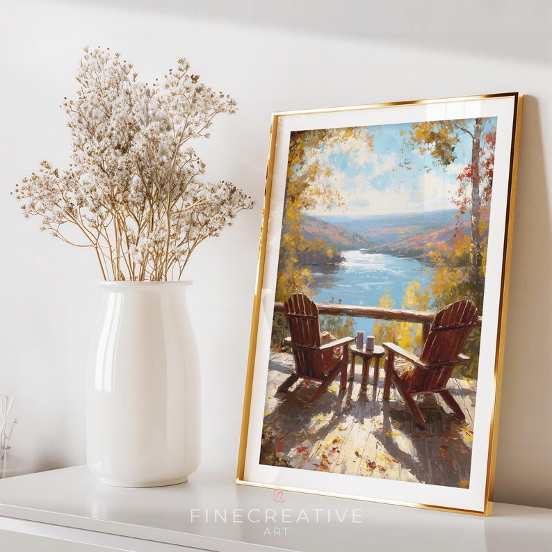 May include: A framed art print featuring a scenic view of a lake and two wooden chairs on a deck. The artwork has a gold frame and depicts an autumn landscape with vibrant colors. A white vase with dried flowers is on the left.