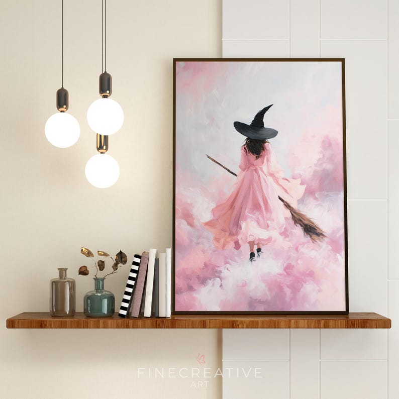 May include: A framed art print depicts a witch in a pink dress and black hat flying on a broom through a pink and white cloudy sky. The artwork is displayed on a wooden shelf with decorative items.