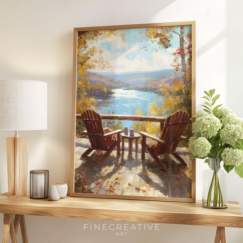 May include: A framed art print depicts a serene lake scene with two wooden Adirondack chairs on a deck. The artwork features a vibrant autumn landscape with colorful trees and a calm blue lake. The print is framed in a light brown wooden frame.