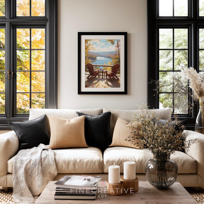 May include: A framed landscape art print depicting two Adirondack chairs on a deck overlooking a lake. The print is displayed above a cream-colored sofa with black and tan pillows. The room has large black framed windows.