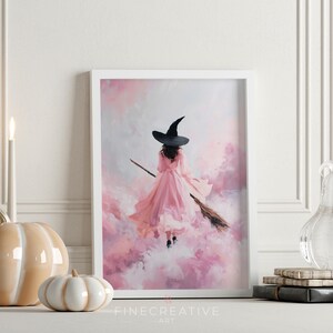May include: A framed art print featuring a witch in a pink dress and black hat flying on a broomstick through a pink and white cloudy sky. The artwork is signed "Fine Creative Art" at the bottom.