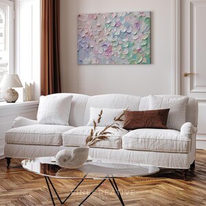 Op de afbeelding: Een woonkamer met een witte bank, kussens en een glazen salontafel. Boven de bank hangt een abstract schilderij met een textuur, bloemblad-achtig ontwerp in pastelkleuren. De kamer is licht en luchtig en heeft een warme, uitnodigende sfeer.