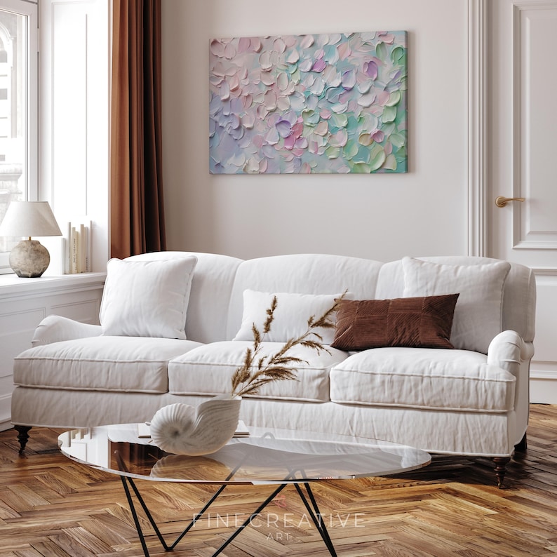 Op de afbeelding: Een woonkamer met een witte bank, kussens en een glazen salontafel. Boven de bank hangt een abstract schilderij met een textuur, bloemblad-achtig ontwerp in pastelkleuren. De kamer is licht en luchtig en heeft een warme, uitnodigende sfeer.
