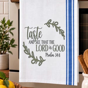 May include: A white kitchen towel featuring a blue vertical striped pattern on the right side. The centre displays the text "Taste and see that the Lord is good Psalm 34:8" in black lettering, framed by two olive green leafy branch illustrations.