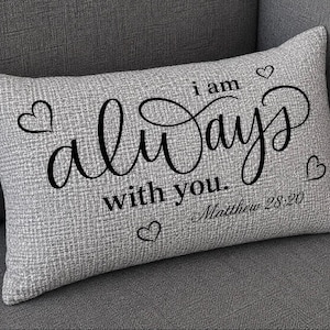 May include: A rectangular grey textured throw pillow featuring black printed text that reads "i am always with you." and "Matthew 28:20". The design includes four small black heart outlines positioned around the central lettering.