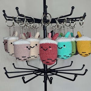 May include: A collection of crocheted boba tea keychain charms in various colors, including pink, cream, red, teal, green, and yellow. Each charm features a white crocheted lid and a cute cartoon face.