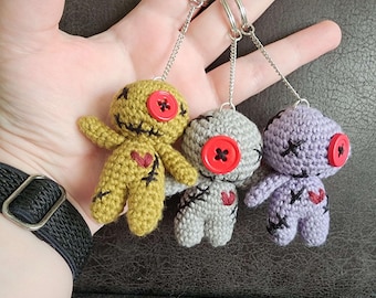 May include: Three small crochet dolls attached to silver metal keychains. The dolls are mustard yellow, light gray, and light purple. Each doll features a single red button eye, black stitched X-shaped eyes and mouths, and a small red heart.