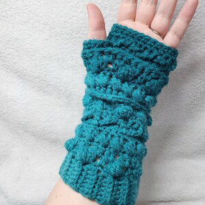 May include: Teal crochet fingerless gloves with a textured, raised pattern. The gloves are made of a soft yarn and are designed to cover the hand and wrist, leaving the fingers free. A handmade accessory.
