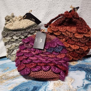 May include: Three handmade crochet drawstring bags in shades of beige, purple, and red. The bags feature a scale-like pattern and a drawstring closure. Each bag has a tag attached. The bags are displayed on a textured surface.