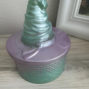 May include: A round container with a lid featuring a sculpted, spiraled, mint-green pointed hat on top. The lid is a metallic lavender color, and the base of the container has a textured, brick-like pattern in a shimmering mint-green finish.