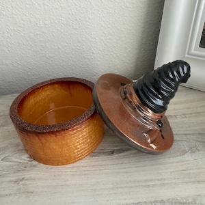 May include: A ceramic jar with a textured, golden-brown brick pattern on the exterior and a smooth, glossy interior. The lid is shaped like a pointed, spiraled black hat with a wide, metallic copper-colored brim that rests beside the jar base.