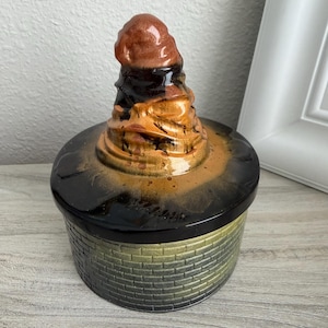 May include: A round ceramic lidded jar featuring a textured brick pattern in shades of green and gold on the base. The lid is glossy black with a central, raised, spiraled sculptural element in shades of brown, tan, and dark chocolate brown.