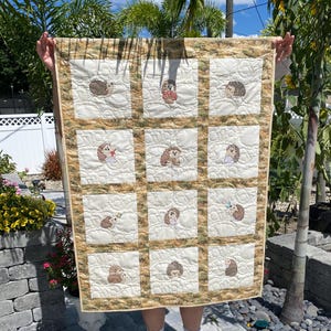 May include: A handmade quilt featuring embroidered hedgehogs in various poses. The quilt has a cream-colored background with a grid pattern and a decorative border. Each square showcases a different hedgehog design, adding a whimsical touch to the textile.