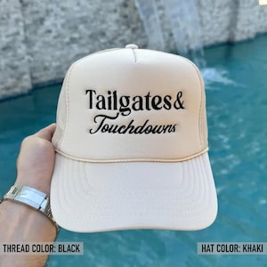 May include: Khaki trucker hat with the text "Tailgates & Touchdowns" embroidered in black. The hat has a white front panel and a mesh back. A stylish accessory for game day.