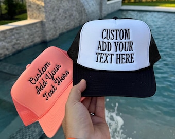 Op de afbeelding: Twee foam trucker petten, een koraalkleurige en een witte met een zwarte mesh achterkant, beide voorzien van zwarte geborduurde tekst Custom Add Your Text Here. De petten hebben een gestructureerd voorpaneel en een gebogen klep.