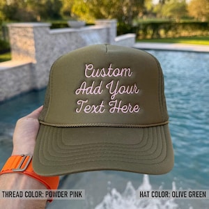 May include: Olive green trucker hat with the text "Custom Add Your Text Here" embroidered in powder pink. The hat features a mesh back and a curved brim. The hat is a stylish accessory.