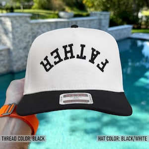 May include: A white and black baseball cap with the word "FATHER" arched across the front in black letters. The hat has a flat brim and a small black button on top. The thread color is black.