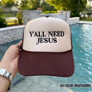 May include: A brown, tan, and brown trucker hat with the text "Y'ALL NEED JESUS" in black. The hat has a mesh back and a curved brim. The hat is being held up in front of a pool.