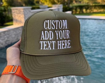 Op de afbeelding: Een olijfgroene truckerpet met een schuimrubberen voorpaneel met de wit geborduurde tekst CUSTOM ADD YOUR TEXT HERE. De pet heeft een bijpassende olijfgroene mesh achterkant, een gestructureerde klep met decoratieve stiksels en een dun koord.