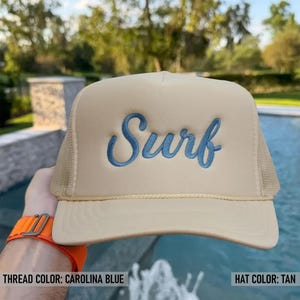 May include: Tan trucker hat with the word "Surf" embroidered in Carolina blue script. The hat features a mesh back and a curved brim. The hat is tan in color.