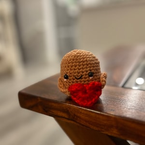 Peut inclure: Une petite figurine en crochet marron avec un visage souriant tenant un cœur rouge. La figurine est posée sur une table en bois.