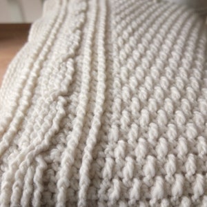 May include: Cream-colored knitted blanket with various textured patterns. The blanket features a combination of cable knit and bobble stitch designs, creating a cozy and tactile appearance. The soft, off-white color adds to its warmth and inviting appeal.