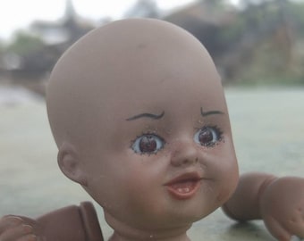 May include: A porcelain doll head with brown skin, painted brown eyes, and a painted smile. The doll head is missing its body and is sitting on a light green surface. Two brown doll arms are also visible in the image.