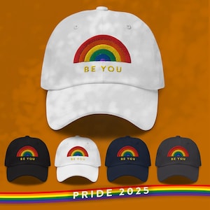 Può includere: Cappellini da baseball bianchi, neri, blu navy e grigi con un arcobaleno e il testo "BE YOU" ricamato sul davanti. I cappellini sono disposti in fila con una striscia arcobaleno sotto e il testo "PRIDE 2025" al centro.