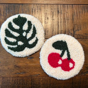 May include: Two round, off-white coasters with a textured, looped surface. One coaster features a dark green monstera leaf design, and the other has a pair of red cherries with green stems. The coasters are placed on a wooden surface.
