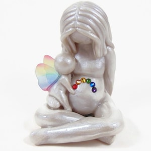 May include: A white ceramic figurine of a woman holding a small fairy with rainbow wings. The woman is sitting with her legs crossed and has a rainbow of rhinestones on her belly.
