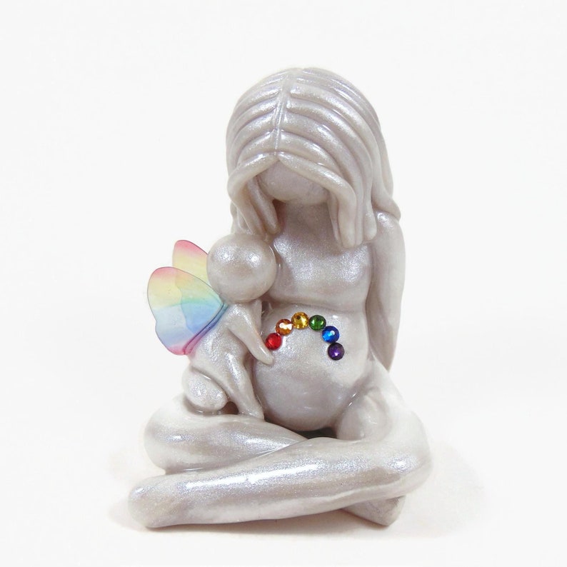 May include: A white ceramic figurine of a woman holding a small fairy with rainbow wings. The woman is sitting with her legs crossed and has a rainbow of rhinestones on her belly.