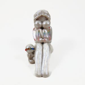 May include: A pearlescent sculpture of a parent figure embracing two smaller figures, with a smaller figure in front. The figures have rainbow accents and colorful details. The sculpture is on a white background.