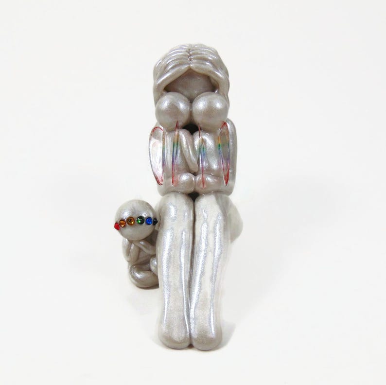 May include: A pearlescent sculpture of a parent figure embracing two smaller figures, with a smaller figure in front. The figures have rainbow accents and colorful details. The sculpture is on a white background.